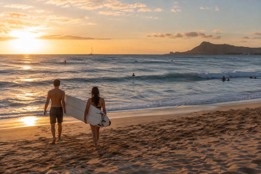 Two people carrying a surfboard across golden sand toward gentle waves at an Oahu beach