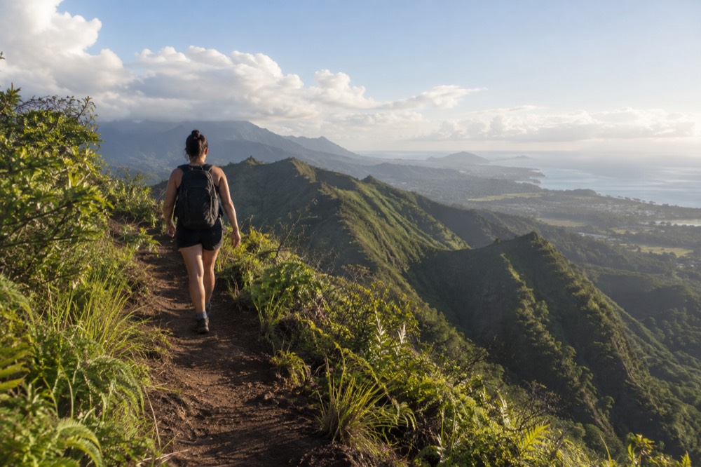 A hiker walking a ridgeline trail above Oahu's green windward valleys in morning light