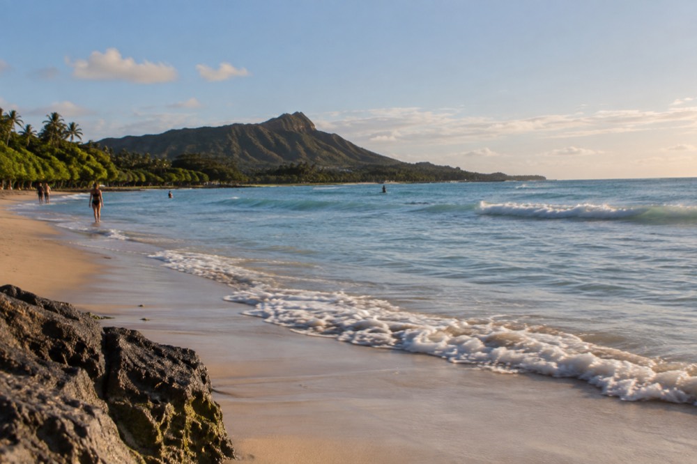 A calm Oahu beach at golden hour with gentle waves and distant green ridgelines
