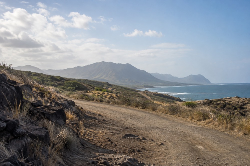 A rough dirt road curving through dry coastal scrub toward distant mountains on Oahu