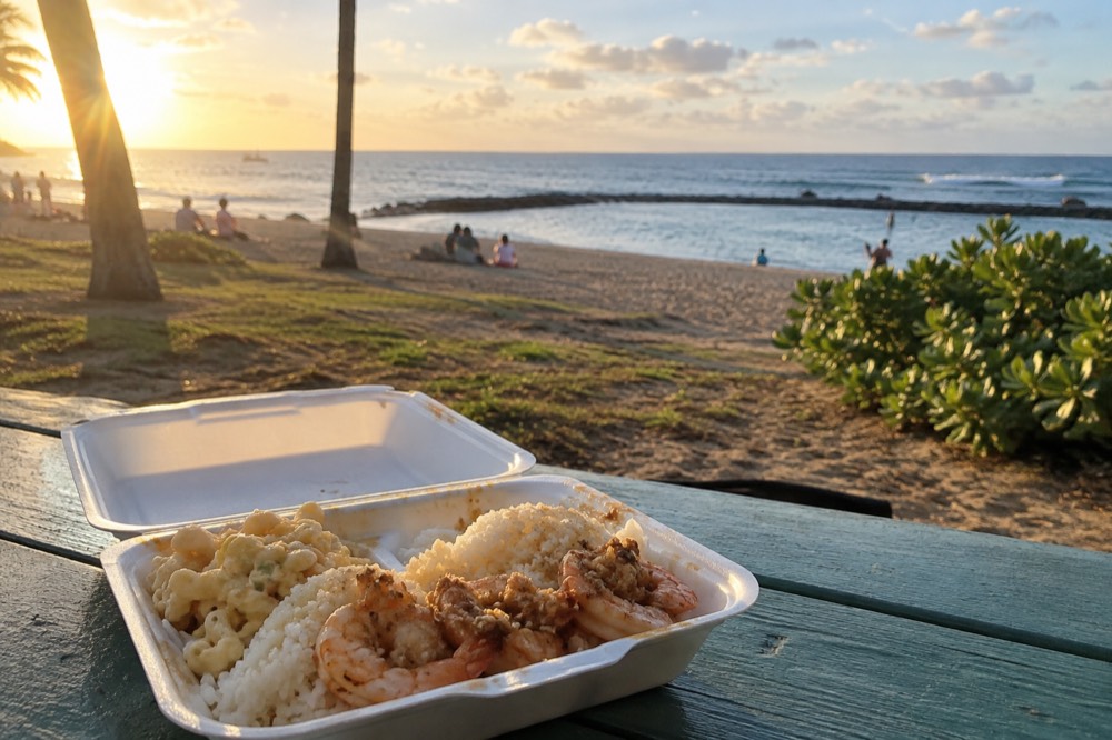 A plate lunch with rice, macaroni salad and garlic shrimp served in a container at a picnic table