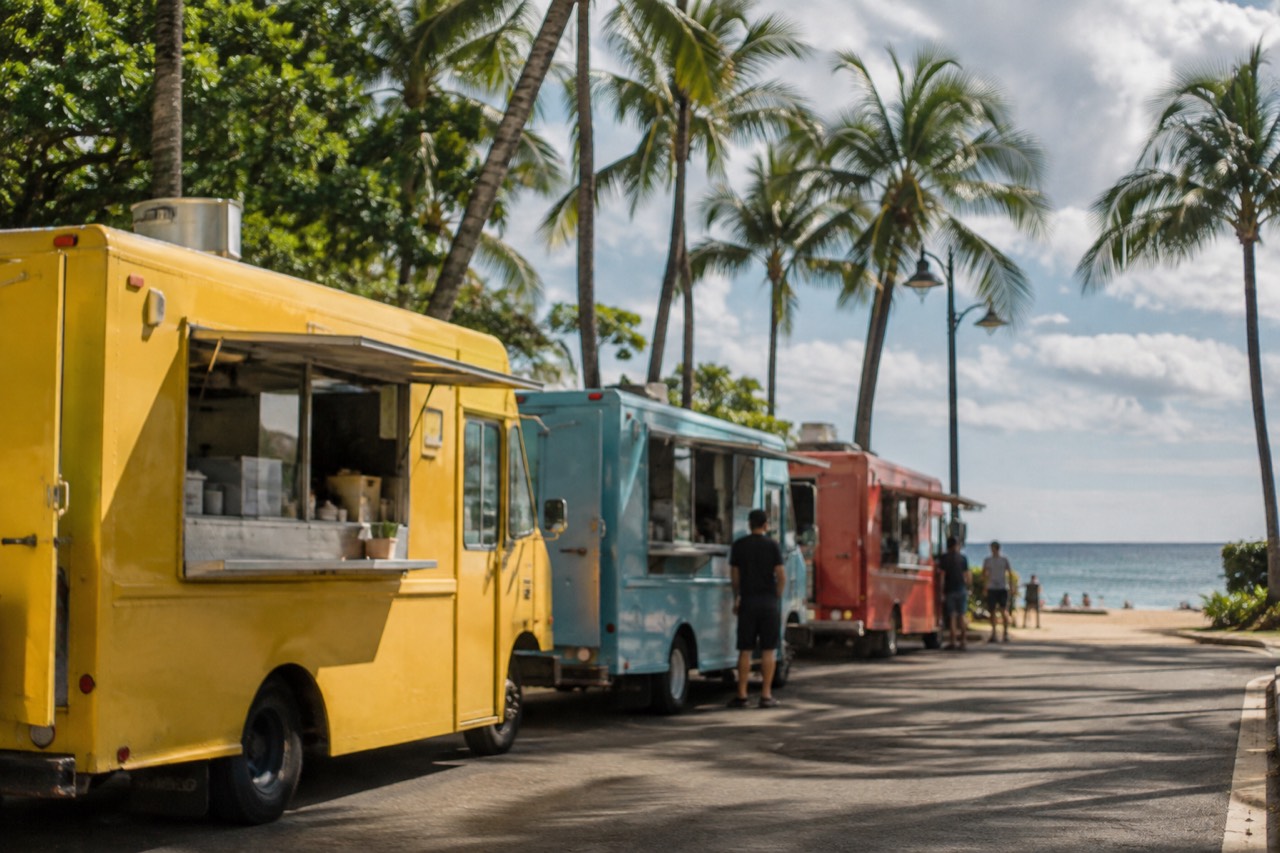 A row of food trucks parked beside a beach road on Oahu with palm trees behind them