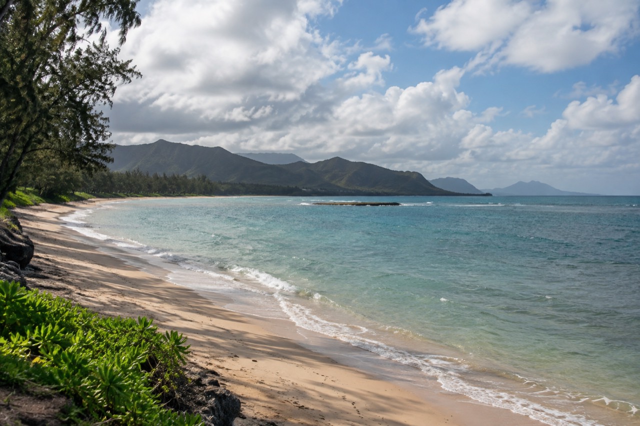 A wide, mostly empty sand beach backed by ironwood trees on the windward coast of Oahu under scattered clouds