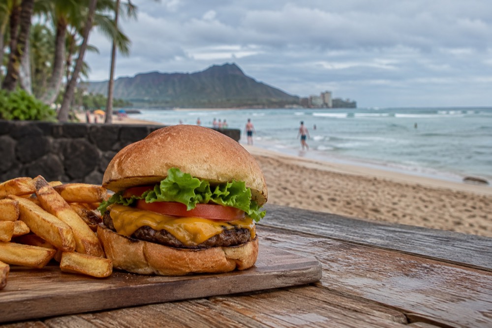 A hamburger with lettuce, tomato and cheese on a wooden board beside a beach view