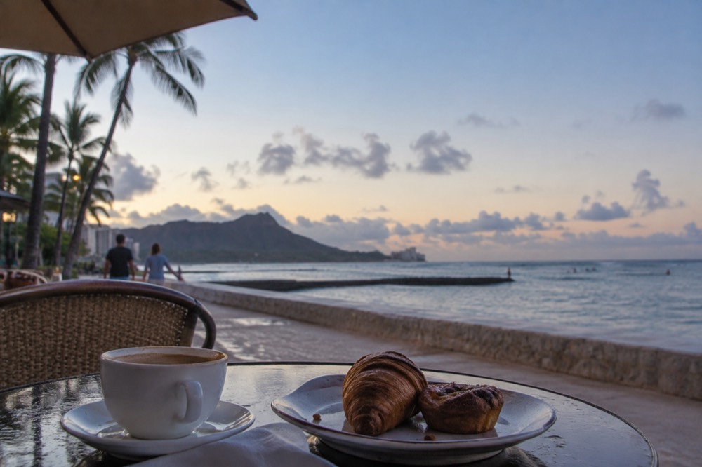 A shaded cafe table on a Honolulu sidewalk with a cup of coffee and a plate of pastries in morning light