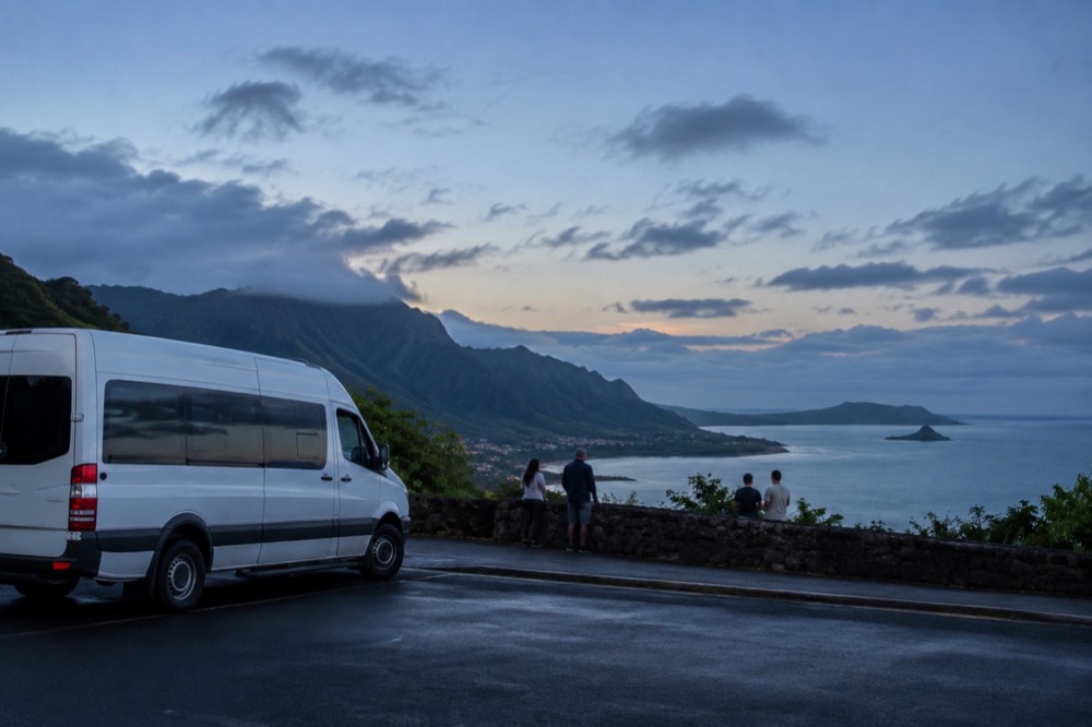 A tour van parked at a roadside lookout above the windward coast of Oahu with green ridges and ocean in the background