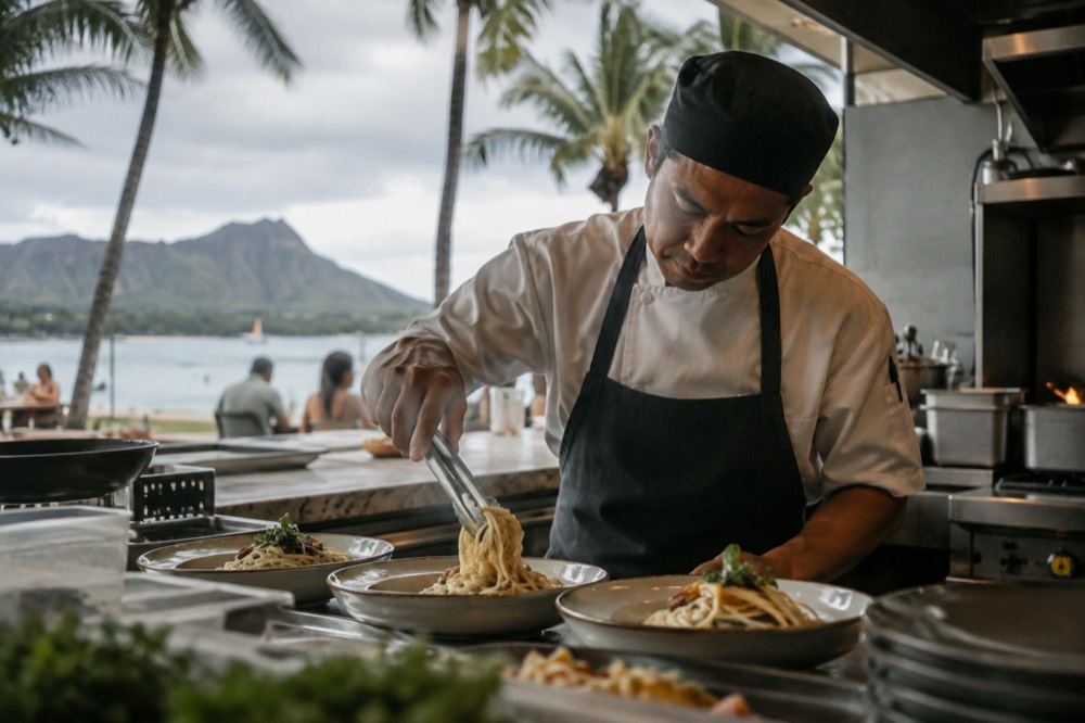 A chef plating fresh pasta in an open restaurant kitchen with a view of palm trees outside