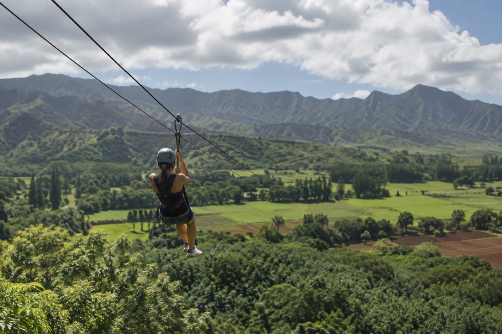A zipline rider in a harness gliding above green ridges and farm fields on the North Shore of Oahu