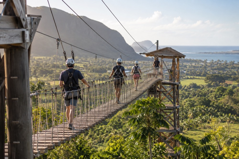 Visitors in helmets and harnesses walking a suspension bridge between zipline platforms above tropical farmland