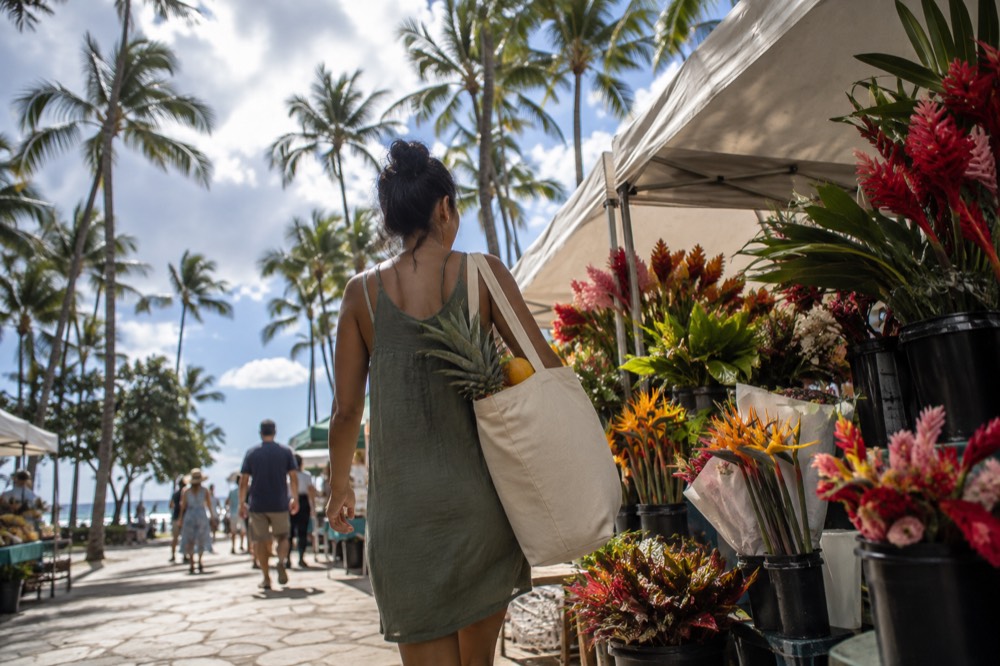 A shopper carrying a canvas bag of tropical fruit past flower stalls at an outdoor market