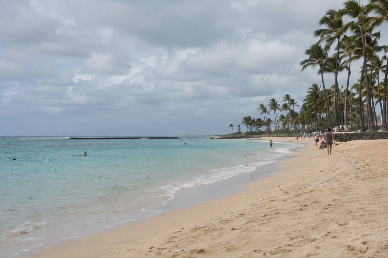 A wide sandy beach in Waikīkī with calm turquoise water and palm trees along the shore