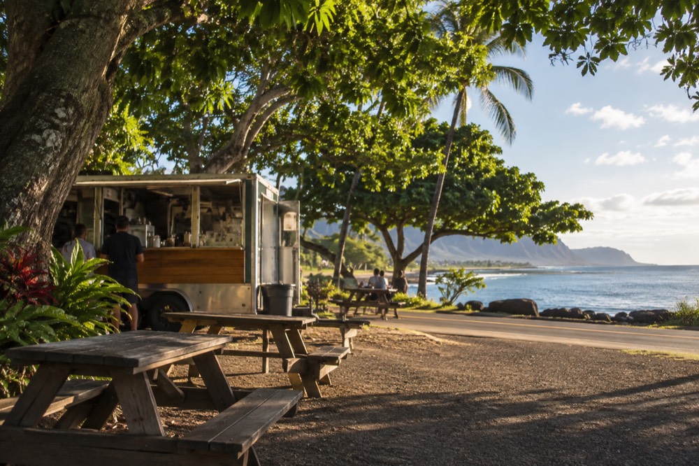 A food truck parked beside a country road on the North Shore of Oahu with picnic tables under trees
