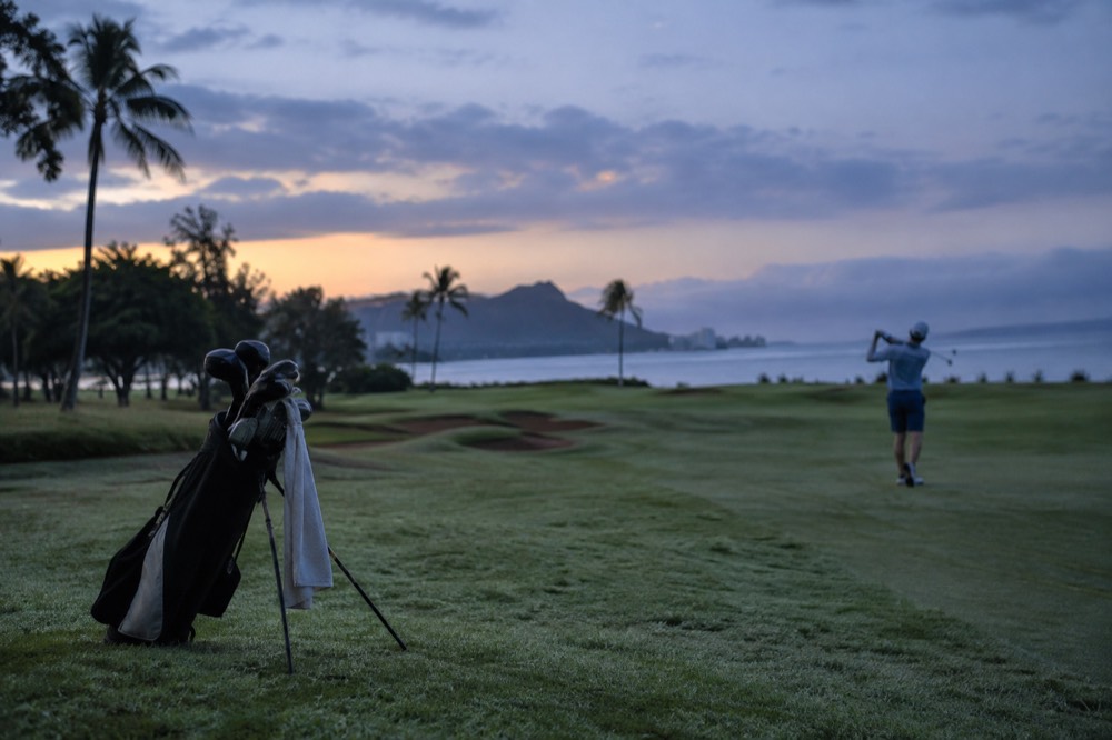 A golfer's bag and clubs resting on dew covered grass beside an Oahu fairway at sunrise