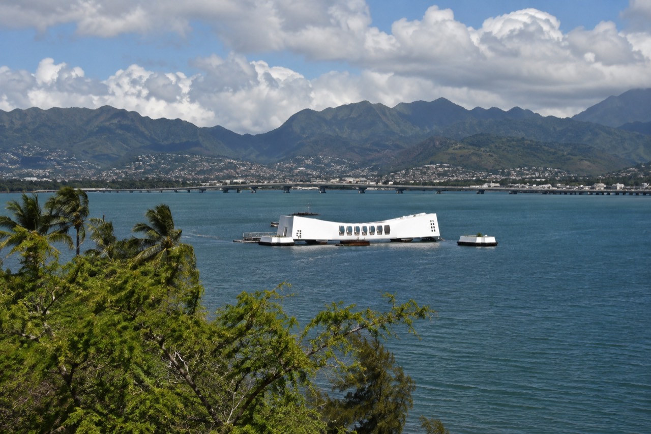The USS Arizona Memorial white structure resting above the water at Pearl Harbor with green mountains behind it