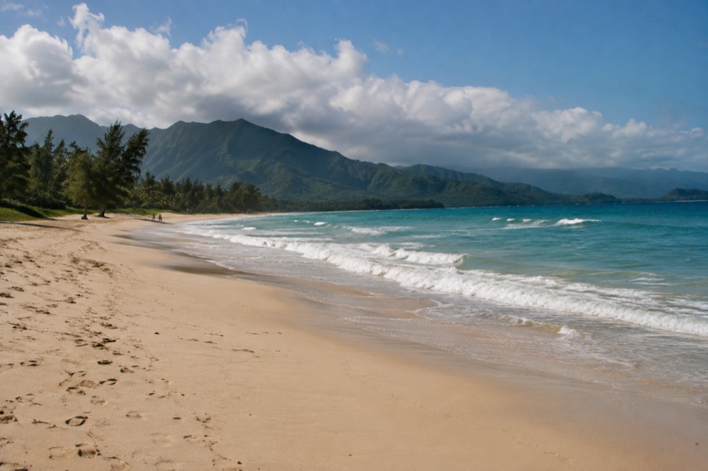 A wide empty beach with soft sand and forested mountains rising behind it under morning clouds