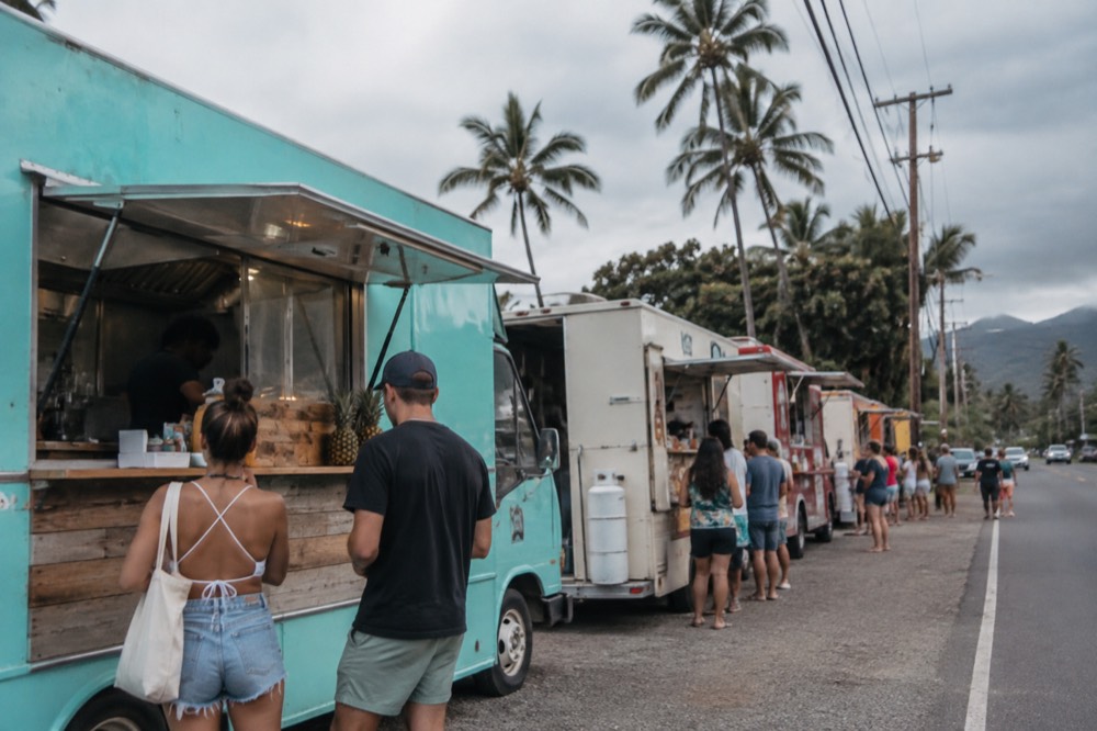 Food trucks parked in a line beside a country road with people waiting at a service window