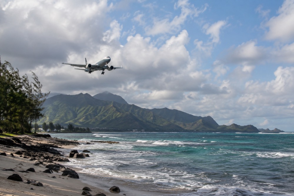 A passenger jet descending over Oʻahu's windward coast with green ridgelines and blue ocean below