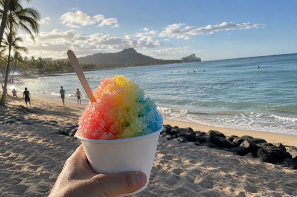 A mound of colorful shave ice in a paper cup held up against a sunny Hawaiian beach backdrop