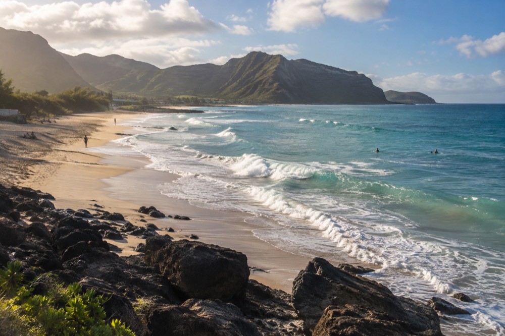 A wide sandy beach on Oahu with turquoise water and green mountains in the background under scattered clouds