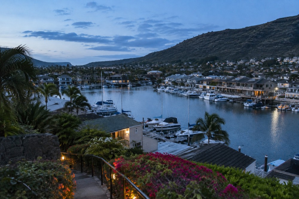 Small boats moored in a calm marina lined with townhouses, hills in the background under a bright sky