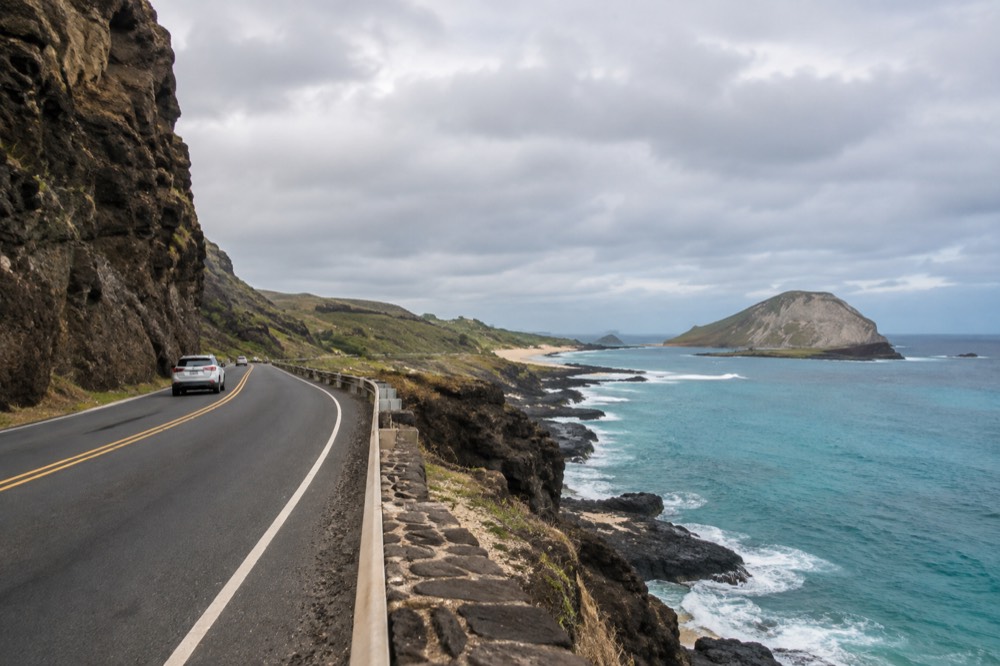 A highway curving along a rocky coastline with turquoise water and an offshore island in the distance
