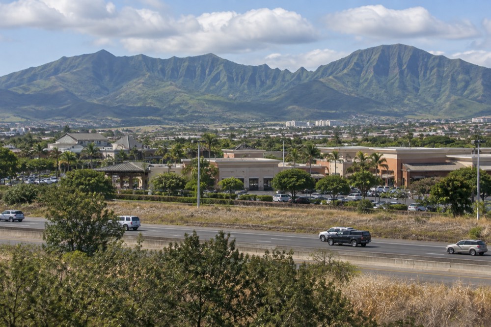 Low-rise buildings and a shopping centre in Kapolei with green ridges behind them under scattered clouds