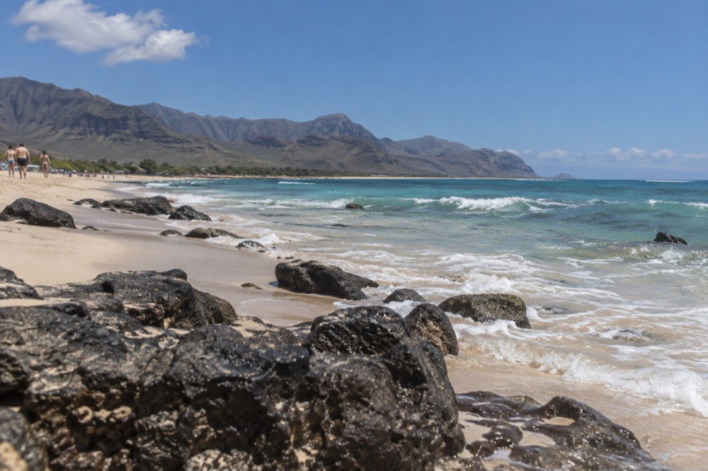 A wide sandy beach on the leeward coast of Oahu under a clear sky, with the Waianae mountains behind