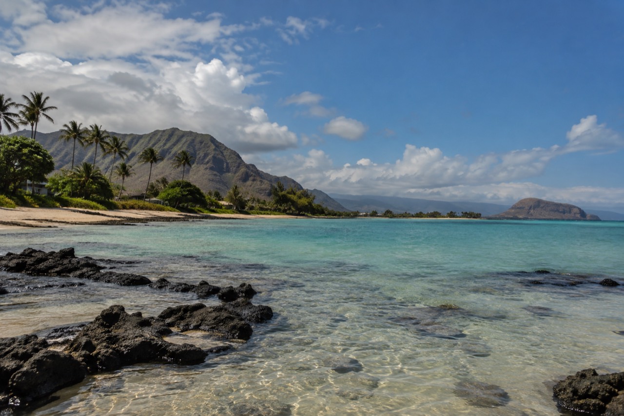 A calm turquoise lagoon along the leeward coast of Oʻahu with palm trees and distant ridges