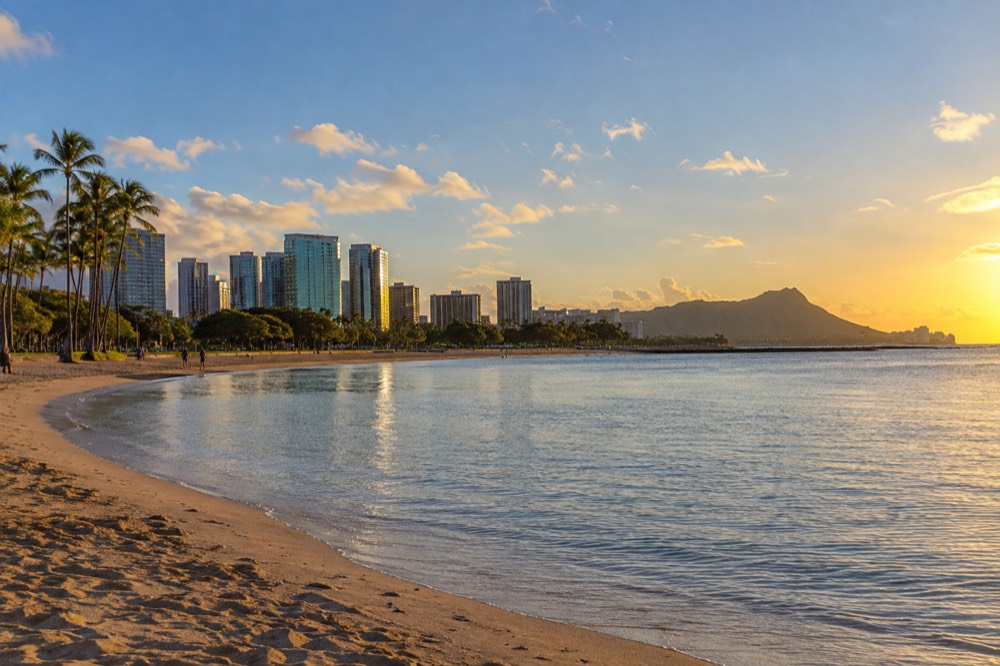 A calm beach lagoon at golden hour with palm trees and distant high-rises