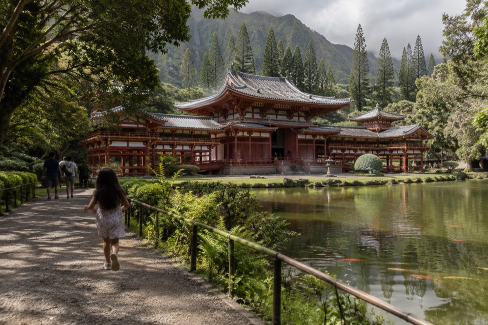 A red Buddhist temple with a green tiled roof standing behind a still reflecting pond with koi