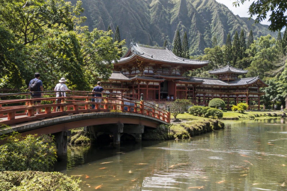 A curved wooden bridge over a pond with koi, leading toward a red temple building under green mountains
