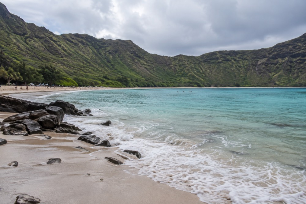 Turquoise water filling a bay ringed by steep green crater walls, with a sandy beach along the shoreline