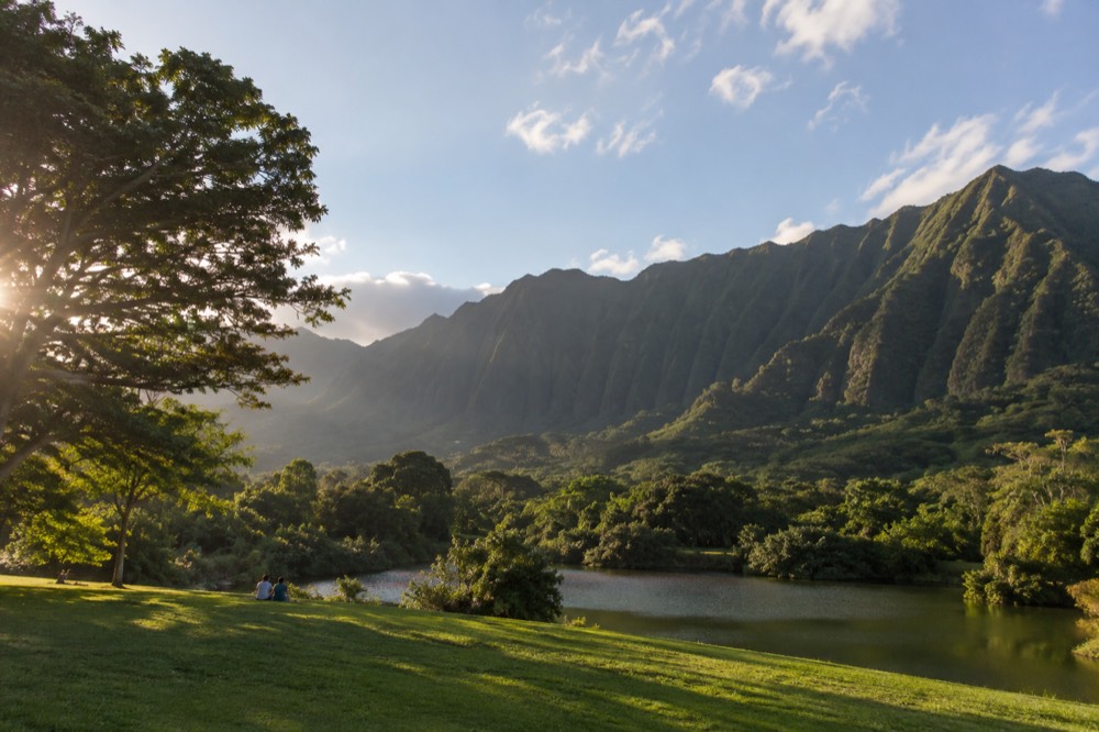 A wide green lawn sloping down to a small lake, with tall fluted volcanic ridges of the Koʻolau mountains rising directly behind the trees.