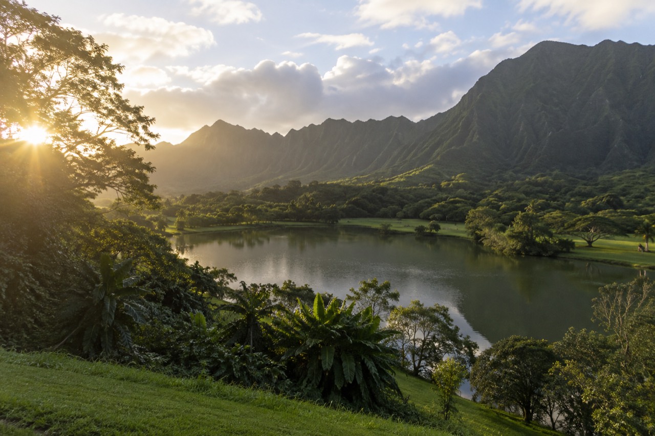 A calm freshwater lake ringed by green lawns, with the steep green cliffs of the Koʻolau mountains rising behind it under scattered clouds.