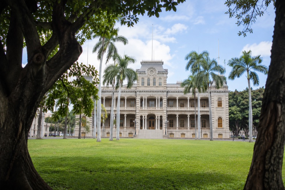 The white facade of ʻIolani Palace fronted by a green lawn and palm trees under a clear Honolulu sky