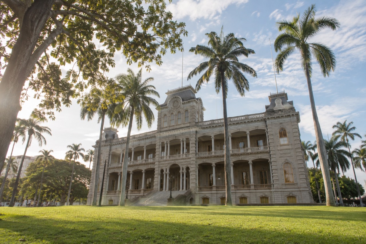 The white facade of ʻIolani Palace fronted by a green lawn and palm trees under a clear Honolulu sky