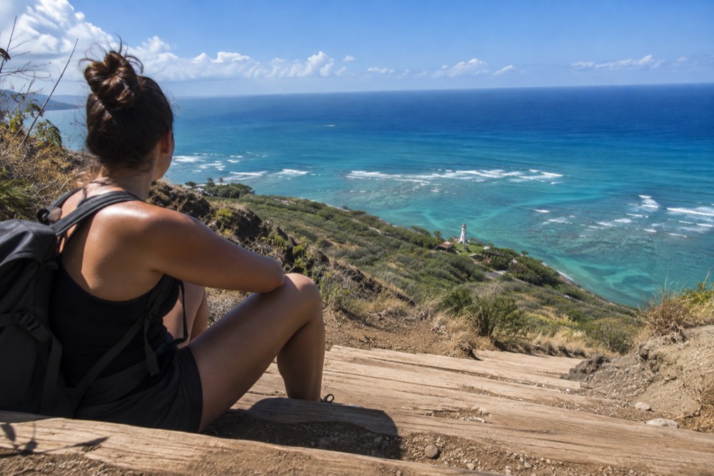 A hiker resting on railway-tie steps high on a crater ridge, with a broad view of a turquoise bay and a distant lighthouse below