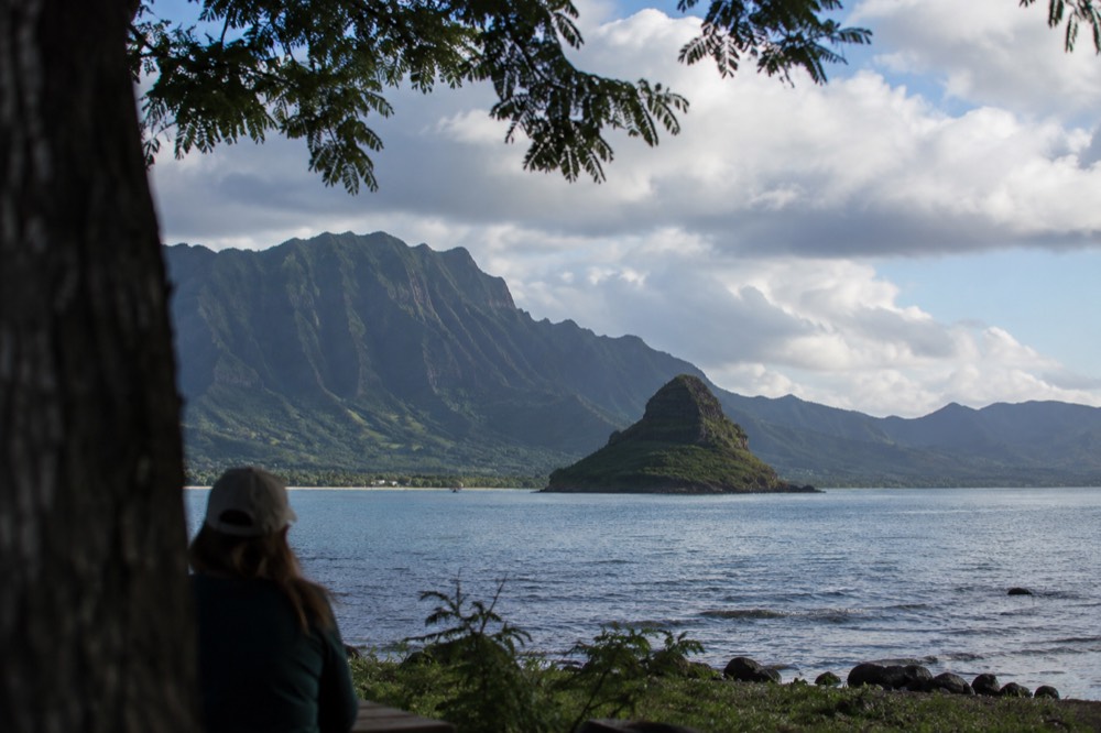A small offshore island in a calm bay with steep green mountain ridges behind it under a partly cloudy sky