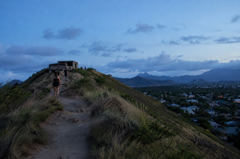 A ridgeline dirt trail above a residential neighbourhood leading toward two small concrete bunkers under a blue sky