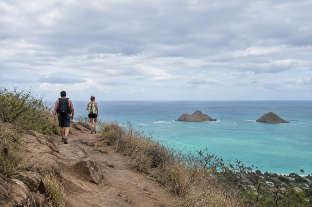 Two hikers on a dirt ridge path with the turquoise ocean and two small offshore islands in the background