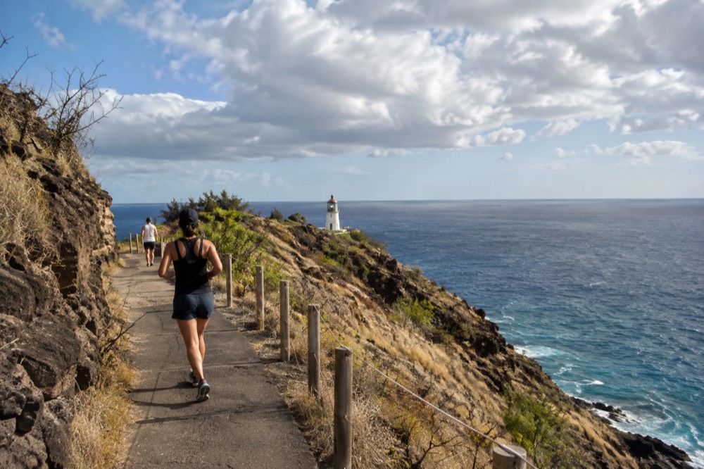 A paved coastal trail climbing a dry hillside toward a white lighthouse on a rocky point above blue ocean