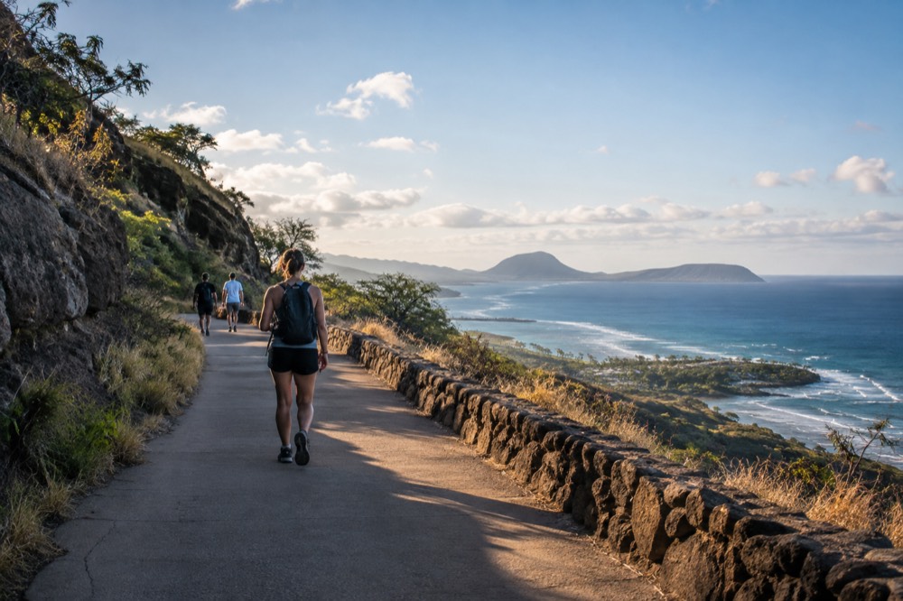 Hikers on a wide paved path near the trailhead with the ocean and offshore islands visible in the distance