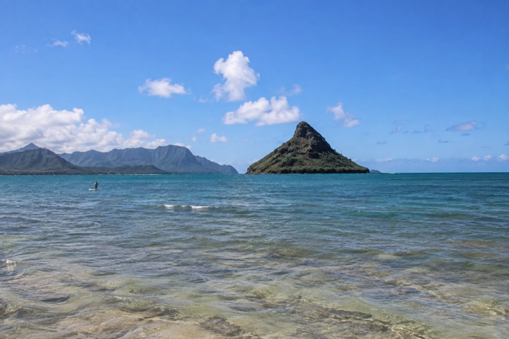 A small volcanic islet rising from calm turquoise water just offshore of a green mountain ridge on a clear morning