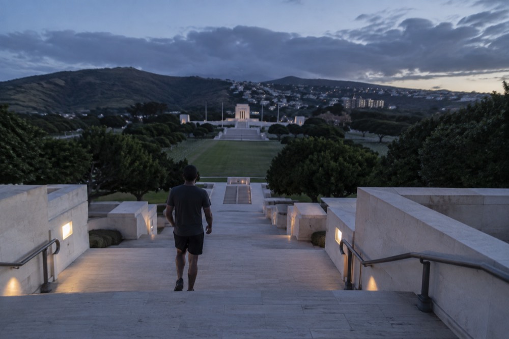 A stone staircase and memorial structure at the edge of a green cemetery crater with mountains beyond