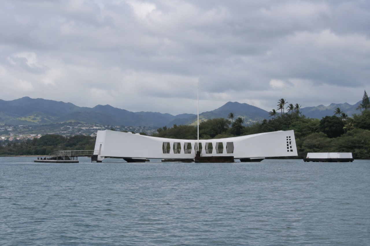 The white USS Arizona Memorial structure resting above the water at Pearl Harbor with green mountains behind it