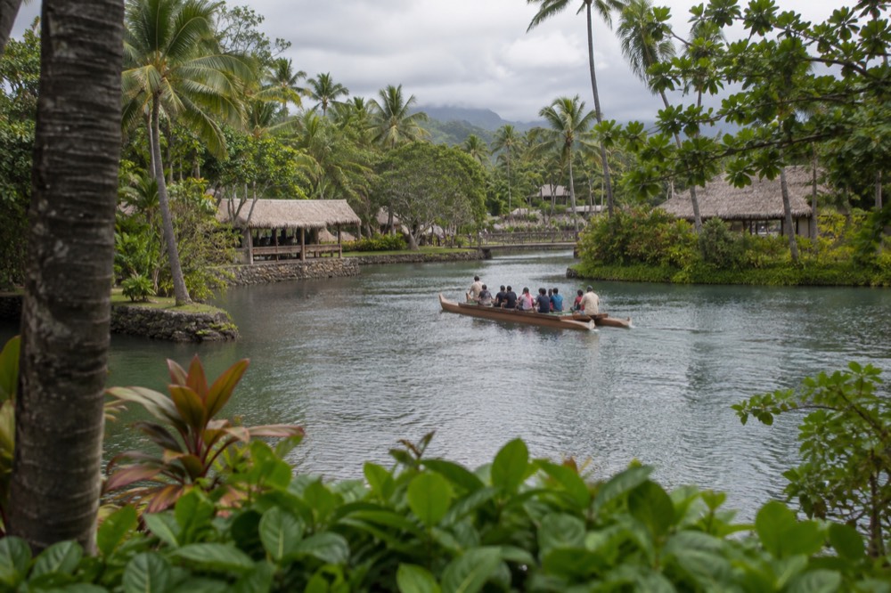 A long double-hulled canoe gliding across a lagoon between green village grounds