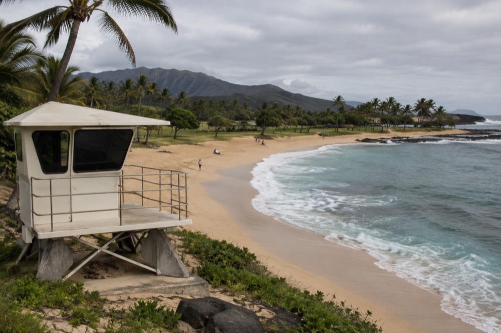 A lifeguard tower above a wide crescent of sand with a green park lawn and palm trees