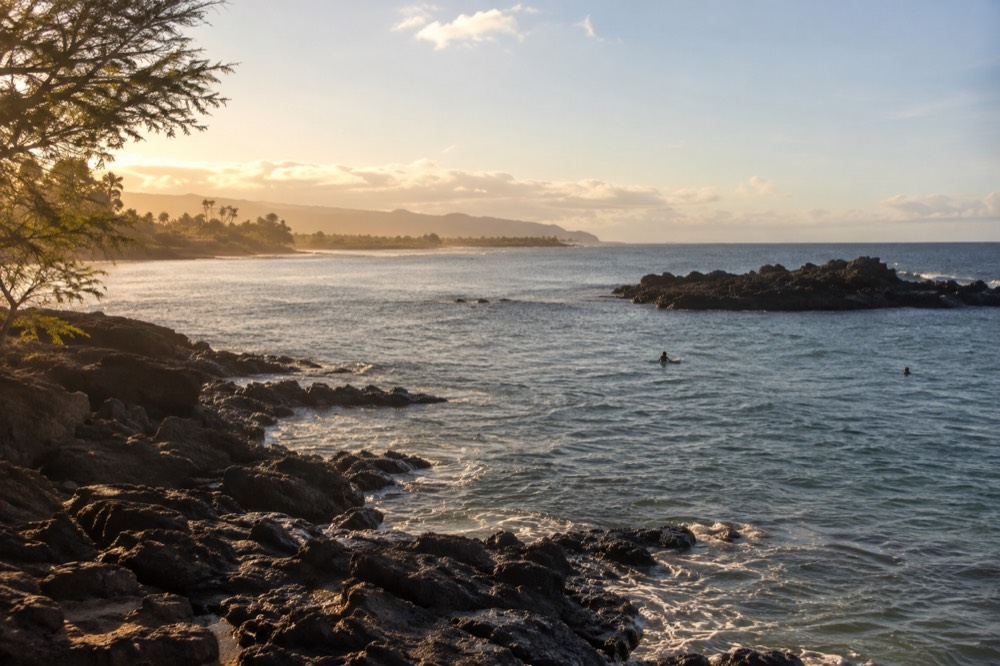 A calm bay at golden hour with a rocky point and distant surf line along the horizon