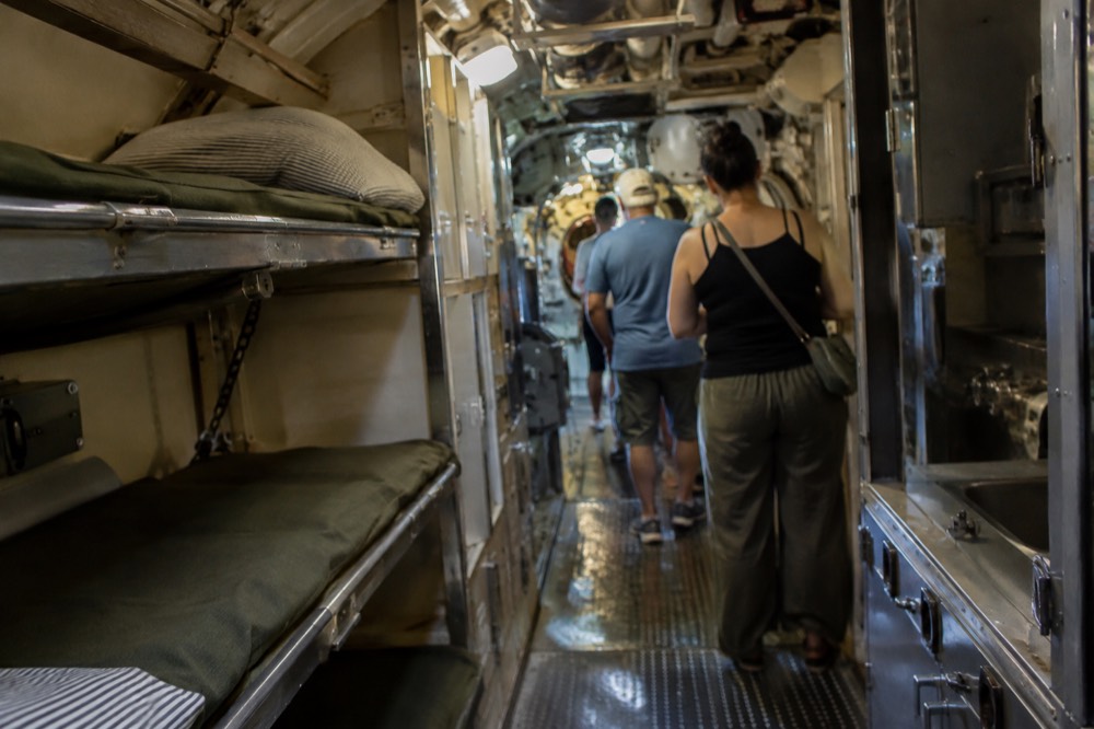 Visitors in a narrow submarine corridor looking at bunks and equipment on both sides