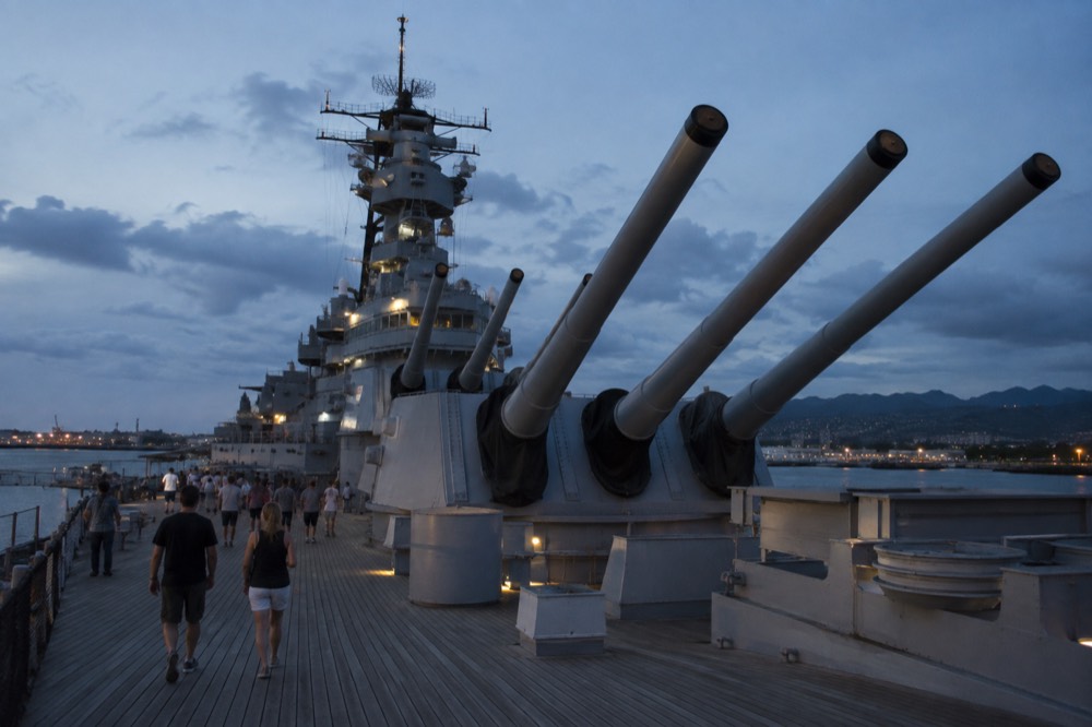 Visitors walking along the wooden deck planking of the battleship Missouri with the ship's large gun turrets in the background